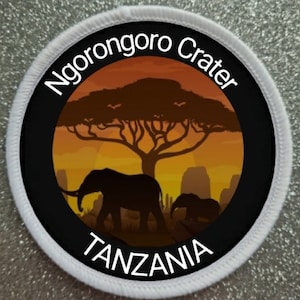 May include: A round patch with a black background and white border. The patch features a silhouette of an acacia tree with two elephants in the foreground. The text "Ngorongoro Crater" is written in white around the top of the patch, and "TANZANIA" is written in white at the bottom.