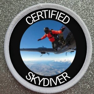 May include: A white round patch with black text that reads "CERTIFIED SKYDIVIER". The centre of the patch features a photo of a skydiver exiting an aeroplane.