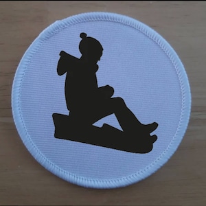 May include: A white round patch with a black silhouette of a person sitting on a sled.