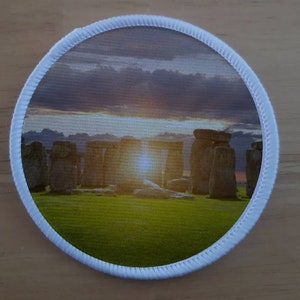 May include: Round patch featuring Stonehenge at sunset. The image shows the ancient monument with a bright sunburst, set against a cloudy sky and green grass. The patch has a white border.