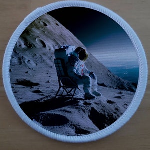 May include: A white astronaut sits on a folding chair on the surface of the moon. The astronaut is wearing a white spacesuit with a blue stripe down the side. The moon's surface is gray and rocky, with craters visible in the background.