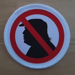 Puede incluir: Un parche de tela blanco con un círculo rojo y una línea roja diagonal que lo atraviesa. Dentro del círculo hay una silueta negra de la cabeza de una persona con un sombrero.