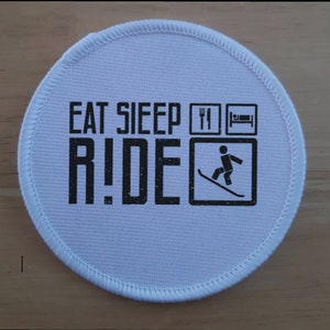 May include: White round patch with black text that reads "Eat Sleep Ride" with a fork and knife, a bed, and a skier on skis in a square.