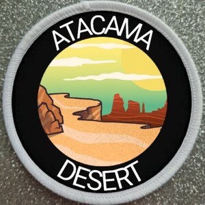 Può includere: Un cerotto circolare bianco e nero con il testo "ATACAMA DESERT" in bianco. Il centro del cerotto presenta un'illustrazione colorata di un paesaggio desertico con formazioni rocciose rosse e un cielo giallo.