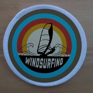 Peut inclure: Un patch rond bordé de blanc avec un design rétro représentant une silhouette d'une personne faisant de la planche à voile contre un coucher de soleil avec un schéma de couleurs bleu, orange et jaune. Le texte "WINDSURFING" est imprimé en blanc sous l'image.