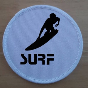 Pode incluir: Patch redondo de tecido branco com uma silhueta preta de uma pessoa surfando e a palavra "SURF" em letras pretas.