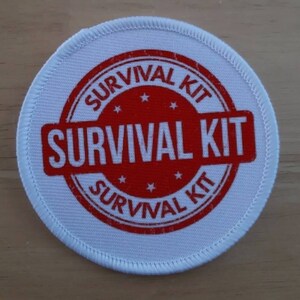 May include: White round fabric patch with a red circle and the text "SURVIVAL KIT" in white letters.