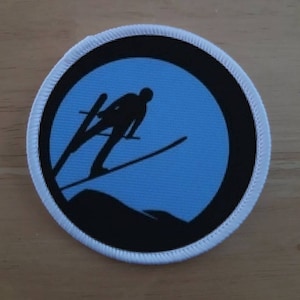 May include: A white and black circular patch with a blue background. The patch features a silhouette of a skier jumping off a ramp.