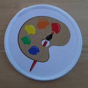 Pode incluir: Um remendo redondo branco com uma paleta de pintura marrom e um pincel. A paleta tem cinco poços de tinta com tinta vermelha, laranja, amarela, verde e azul.