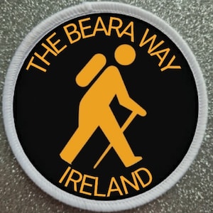 Op de afbeelding: Een ronde zwarte en witte patch met een witte rand. De patch toont een gele stick figure wandelaar met een rugzak en wandelstok. De tekst "THE BEARA WAY IRELAND" staat in het geel rond de wandelaar.