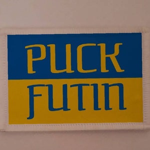 May include: A white patch with blue and yellow stripes. The text "PUCK FUTIN" is written in blue letters on the patch.