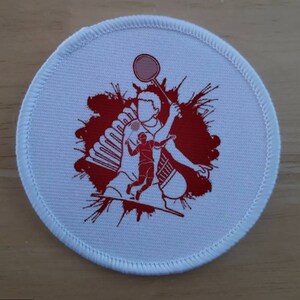 May include: White round patch with red badminton player silhouette and a red splatter background. The player is holding a badminton racket and a shuttlecock.