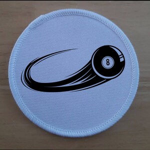 Puede incluir: Parche redondo de tela blanca con un gráfico de bola negra número 8. La bola 8 está en movimiento, con una raya negra detrás.