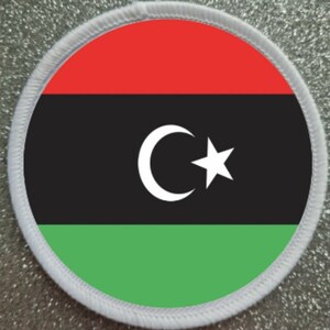 May include: A round, white fabric patch with a red, black, and green Libyan flag design. The flag features a white crescent moon and star on a black background.