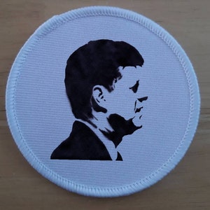 Può includere: Un cerotto rotondo in tessuto bianco con una silhouette nera di una testa e spalle di una persona. La persona è rivolta a destra e ha i capelli corti.