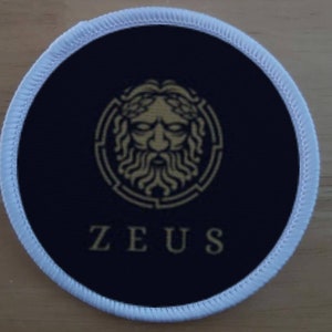 May include: A round, white-edged patch with a black background. A gold-colored image of a face with a beard and a wreath is surrounded by a gold-colored circle. The text "ZEUS" is written in gold letters below the image.