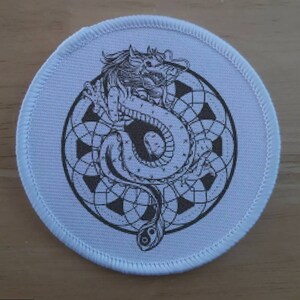 Può includere: Un cerotto di tessuto bianco con un'illustrazione in bianco e nero di un drago avvolto attorno a un simbolo yin yang. Il drago è circondato da un motivo circolare di forme geometriche.