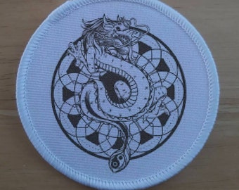 Può includere: Un cerotto di tessuto bianco con un'illustrazione in bianco e nero di un drago avvolto attorno a un simbolo yin yang. Il drago è circondato da un motivo circolare di forme geometriche.