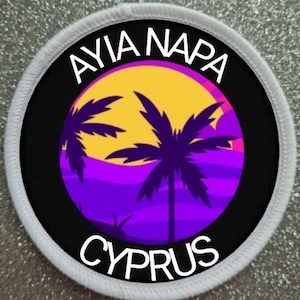 May include: A black and white circular patch with the text "Ayia Napa Cyprus" in white. The patch features a purple and yellow sunset with two palm trees.