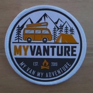 May include: A white, round patch with a black border and the text "MY VANTURE" in yellow letters. The patch also features a yellow camper van, a yellow tent, and a campfire graphic. The text "MY VAN MY ADVENTURE" is written in black around the bottom of the patch, with "EST. 2019" in smaller black text above the campfire.