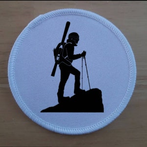 May include: A white round patch with a black silhouette of a person skiing. The person is wearing a helmet and carrying skis on their back. The person is standing on a mountain.