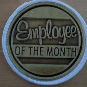 Op de afbeelding: Een ronde witte en gouden patch met de tekst "Employee of the Month" in zwarte letters.
