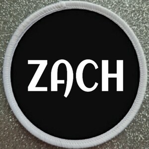 Puede incluir: Parche redondo con centro negro y texto blanco "ZACH". El parche tiene un borde blanco con un borde cosido. El fondo es plateado brillante.