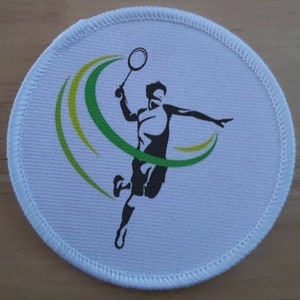 May include: White round fabric patch with a green and yellow design featuring a silhouette of a badminton player with a racket.