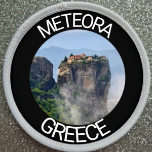 May include: A round white patch with a black circle in the center. The circle contains a photo of a monastery built on a cliff in Meteora, Greece. The text "METEORA" is written in white above the photo and "GREECE" is written in white below the photo.