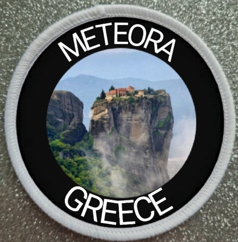 May include: A round white patch with a black circle in the center. The circle contains a photo of a monastery built on a cliff in Meteora, Greece. The text "METEORA" is written in white above the photo and "GREECE" is written in white below the photo.