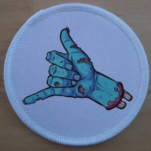 May include: A circular patch with a light blue background and a white stitched border. It depicts a cartoon zombie hand in a shaka gesture, with blue skin, red wounds, and exposed bone.