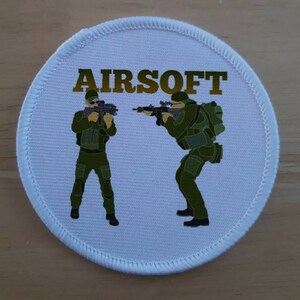 以下が含まれることがあります： 金色の「AIRSOFT」の文字と、迷彩服を着てライフルを持った漫画のキャラクター2人が描かれた白い丸いパッチ