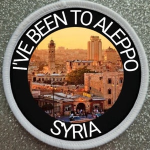 May include: A round, white patch with a black circle in the center. The circle has the text "I've Been To Aleppo Syria" in white lettering around a colorful image of a city skyline.