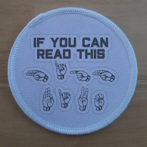 May include: A circular white embroidered patch with a stitched border. The text IF YOU CAN READ THIS is printed in dark gray. Below the text are eight black line-art illustrations of hands forming letters in American Sign Language.