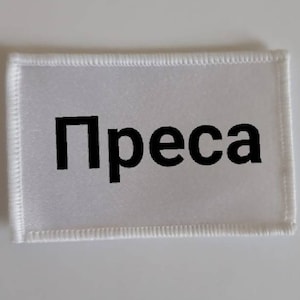 May include: White rectangular patch with black stitching around the edges. The word "Prensa" is printed in black letters on the patch.