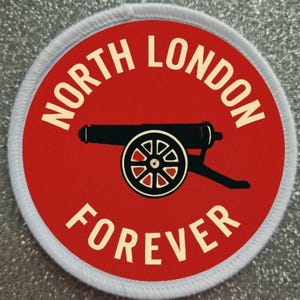 Könnte beinhalten: Ein runder Aufnäher mit rotem Hintergrund und weißem, genähtem Rand. Der Aufnäher zeigt die weißen Worte "NORTH LONDON FOREVER" um eine schwarze Kanonengrafik.