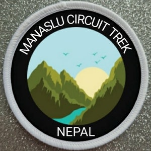 Puede incluir: Parche redondo blanco con borde negro y texto blanco que dice "Manaslu Circuit Trek Nepal". El centro del parche presenta una cordillera verde con un río azul y un sol amarillo.