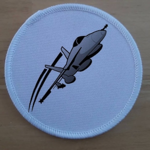 Op de afbeelding: Ronde witte stoffen patch met een grijs silhouet van een straaljager in de vlucht.