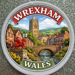 Puede incluir: Parche redondo con una ilustración detallada de Wrexham, Gales. El diseño incluye un dragón rojo, un puente de piedra, una iglesia y los textos "WREXHAM" y "WALES". El parche tiene un borde blanco.