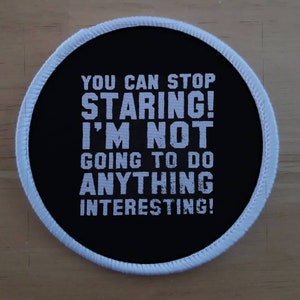 Puede incluir: Un parche redondo negro y blanco con costuras blancas alrededor del borde. El texto en el parche dice "You can stop staring! I'm not going to do anything interesting!"