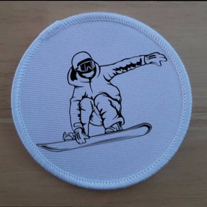 May include: A white round patch with a black and white illustration of a person snowboarding. The person is wearing a helmet and goggles.