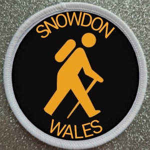 May include: A black and white round patch with a white border. The patch features a yellow stick figure hiker with a backpack and walking stick. The text "SNOWDON WALES" is written in yellow around the hiker.