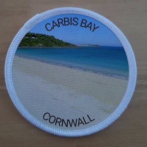 May include: White round patch with a photo of a beach with blue water and white sand. The text "CARBIS BAY CORNWALL" is printed on the patch.