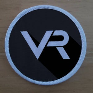 Op de afbeelding: Een witte geborduurde patch met een zwarte achtergrond en de letters "VR" in wit.