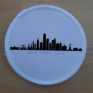 May include: A white round fabric patch featuring a black silhouette of the New York City skyline, including the Empire State Building, the Chrysler Building, and the Statue of Liberty. The text "NEW YORK CITY" is printed below the skyline.
