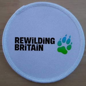 May include: White round patch with black text that reads "Rewilding Britain" and a green, blue, and light blue paw print.
