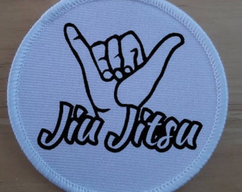 May include: White round patch with a black outline. The patch features a black hand making the shaka sign and the text "Jiu Jitsu" in black.