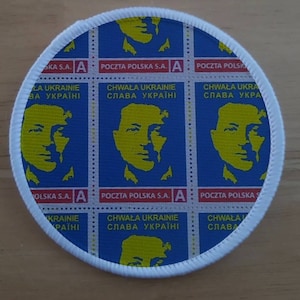 May include: A round white patch with a blue background and a repeated image of a yellow face with the text "CHWAŁA UKRAINIE SŁAWA UKRAINIE" in red and black letters.
