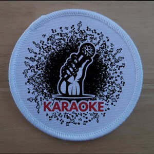 May include: White round patch with a black and white illustration of a hand holding a microphone with musical notes surrounding it. The text "Karaoke" is in red letters.