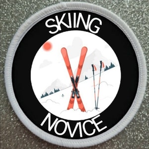 May include: A circular patch with the text "SKIING NOVICE" in white. The design includes orange skis, ski poles, a sun, and a snowy mountain landscape, set against a black background with a white border.